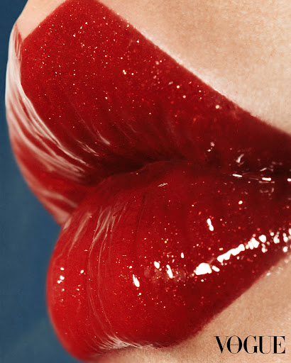 Close-up of a woman's lips covered in glossy red lipstick, set against a solid blue background, with the Vogue logo in the corner.