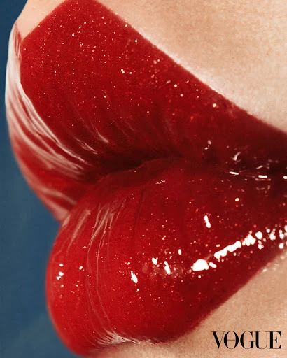 Close-up of a woman's lips covered in glossy red lipstick, set against a solid blue background, with the Vogue logo in the corner.