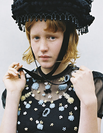 Close-up portrait of a woman with red hair, wearing an elaborate black bonnet and a sheer black dress with floral embroidery.