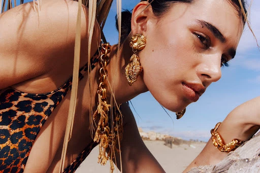Close-up of a woman with gold jewelry posing on a sunny beach, showcasing a fashionable leopard-print swimsuit and tropical plants.