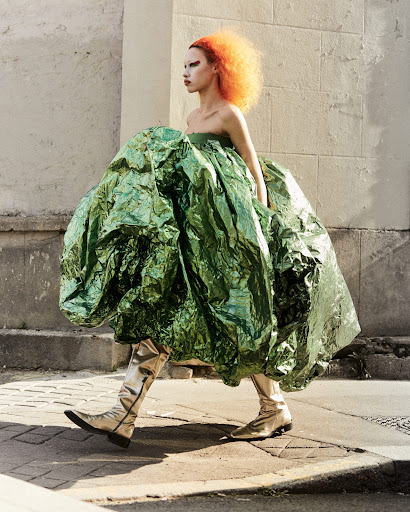 Woman with vibrant orange hair and silver boots walks outdoors, wearing a voluminous, shimmering green dress in a fashion editorial.