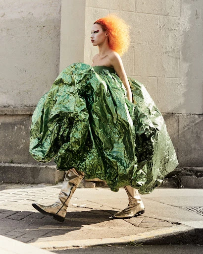 Woman with vibrant orange hair and silver boots walks outdoors, wearing a voluminous, shimmering green dress in a fashion editorial.
