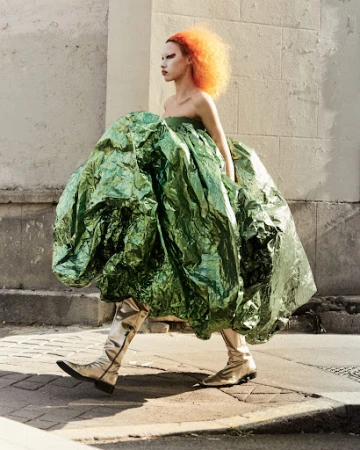 Woman with vibrant orange hair and silver boots walks outdoors, wearing a voluminous, shimmering green dress in a fashion editorial.
