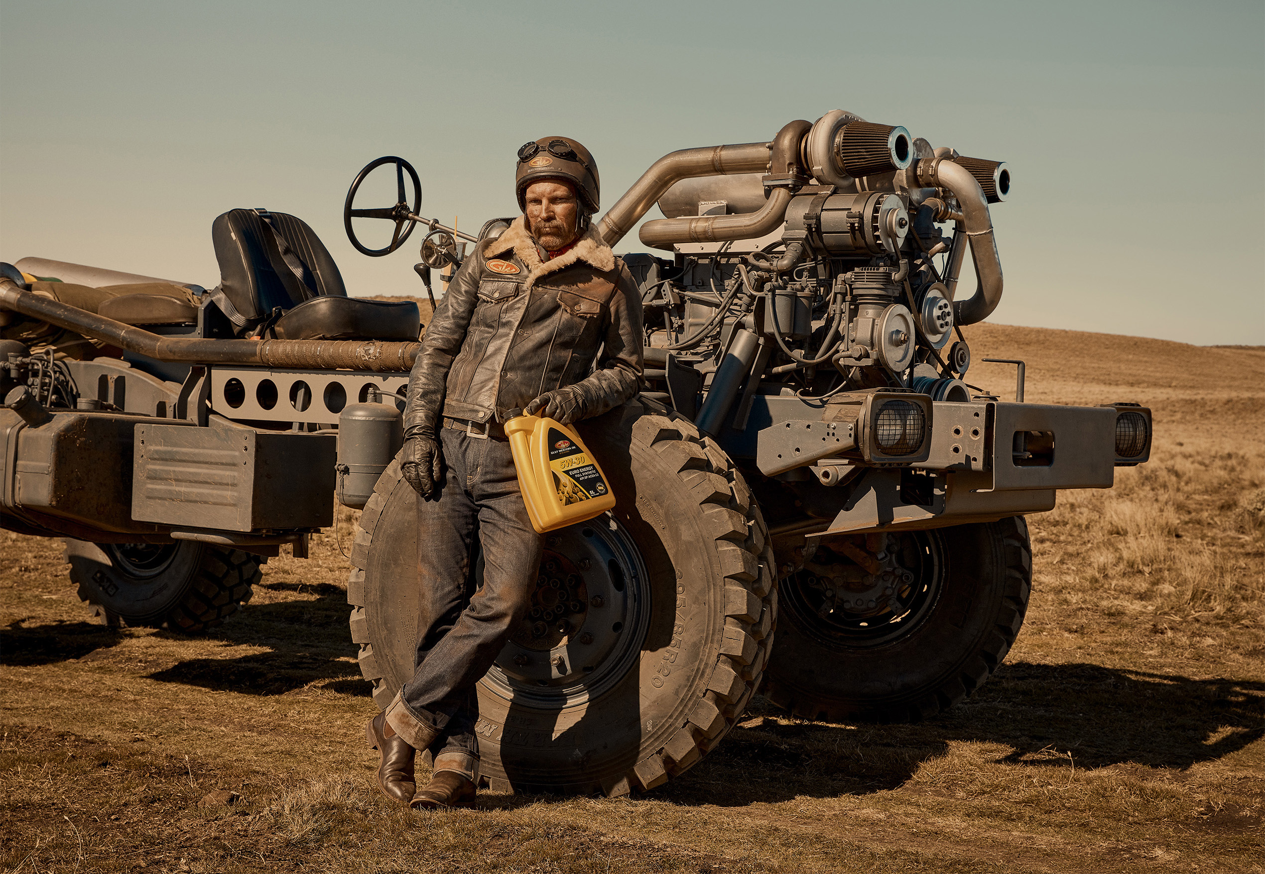 Adult man in driving goggles and leather jacket leans against custom Mad Max-style vehicle in desert landscape, holding motor oil.