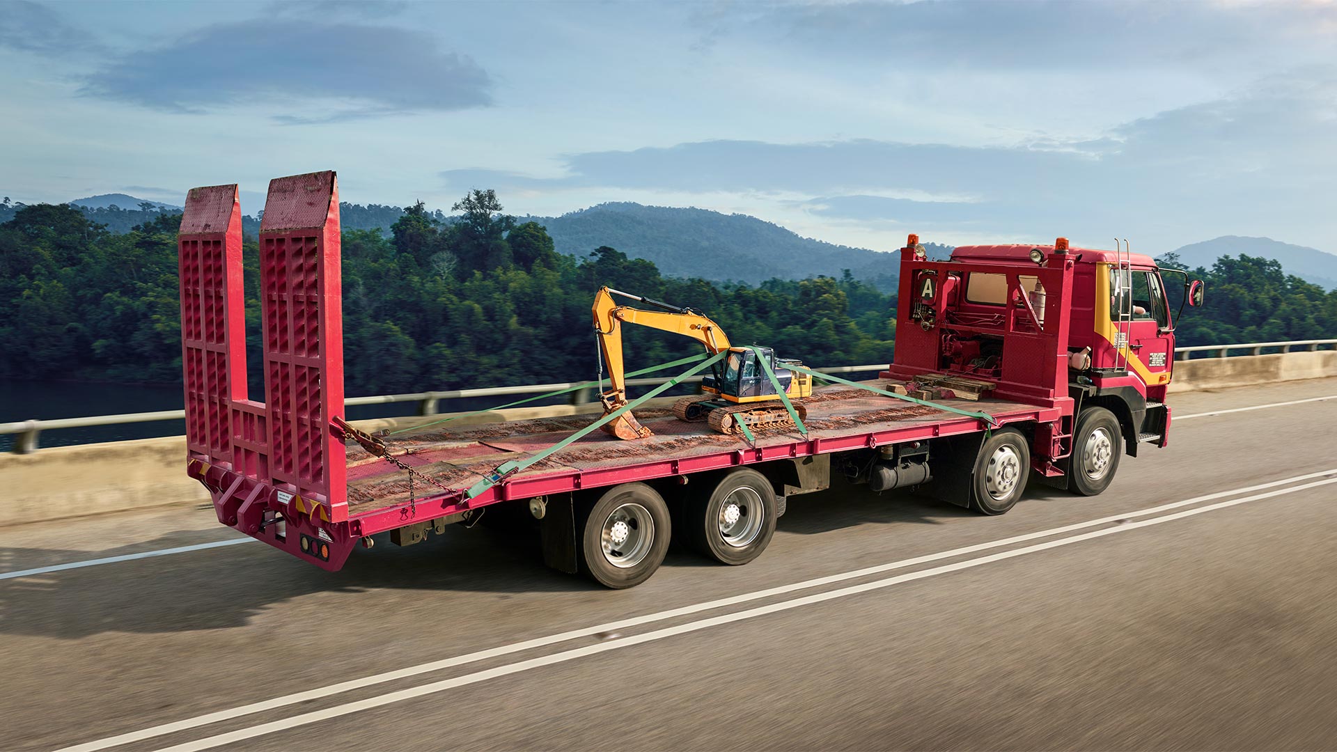 A red flatbed truck transports a yellow excavator down a road. Forest and mountains create a scenic background under a blue, cloudy sky.