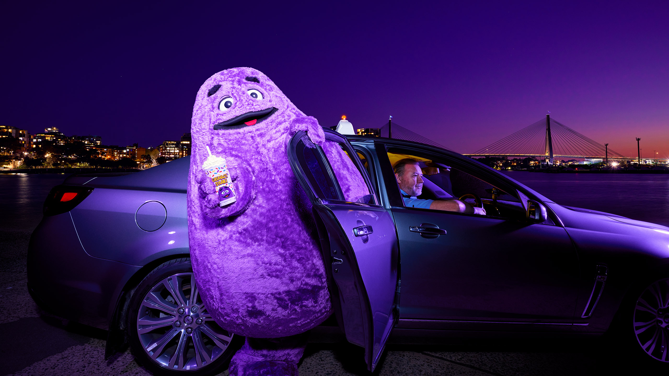 Grimace character in costume holds shake beside car with driver in urban waterfront setting at night; bridge visible.