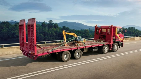 Red flatbed truck transporting a small yellow excavator on a highway bridge surrounded by lush green hills