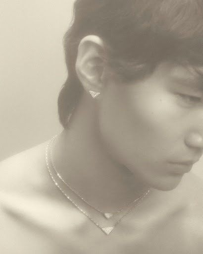 Close-up of a young adult with triangular diamond jewelry: necklace and earrings, soft lighting, pale skin, and a neutral expression.