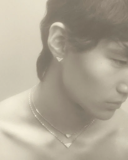 Close-up of a young adult with triangular diamond jewelry: necklace and earrings, soft lighting, pale skin, and a neutral expression.
