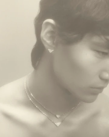 Close-up of a young adult with triangular diamond jewelry: necklace and earrings, soft lighting, pale skin, and a neutral expression.