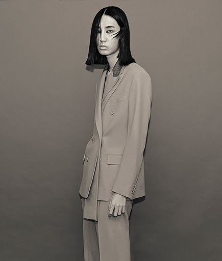 Studio shot of a young adult female with short dark hair in a double-breasted suit, posing against a neutral grey backdrop in diffused light.