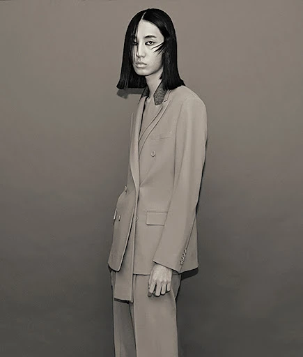 Studio shot of a young adult female with short dark hair in a double-breasted suit, posing against a neutral grey backdrop in diffused light.