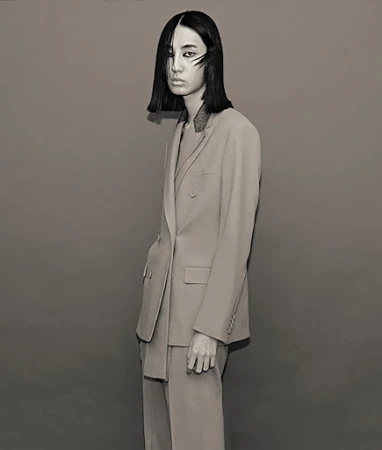 Studio shot of a young adult female with short dark hair in a double-breasted suit, posing against a neutral grey backdrop in diffused light.