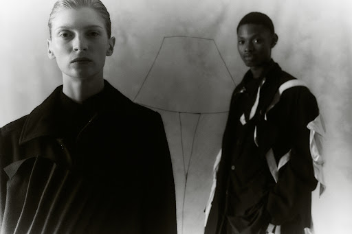 Black-and-white fashion studio shot of two models wearing jackets against a sketched backdrop; one is blond and the other is Black.