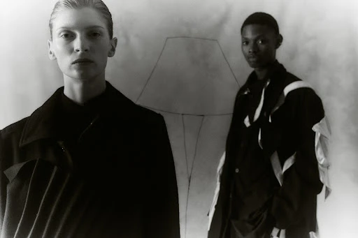 Black-and-white fashion studio shot of two models wearing jackets against a sketched backdrop; one is blond and the other is Black.