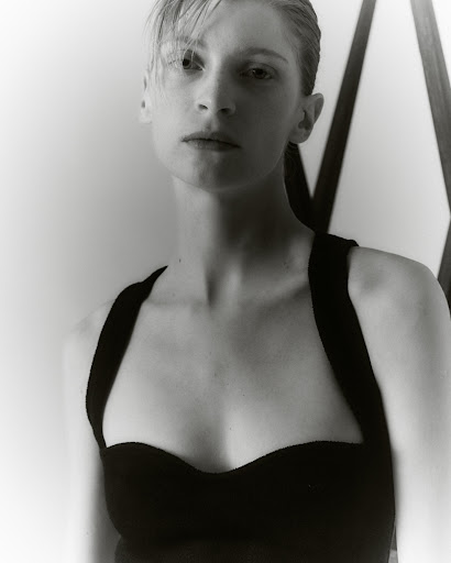 Portrait of a woman with wet-look hair wearing a black top against a light wall and geometric frame.
