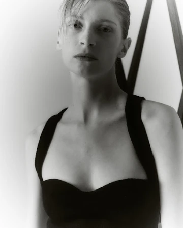Portrait of a woman with wet-look hair wearing a black top against a light wall and geometric frame.
