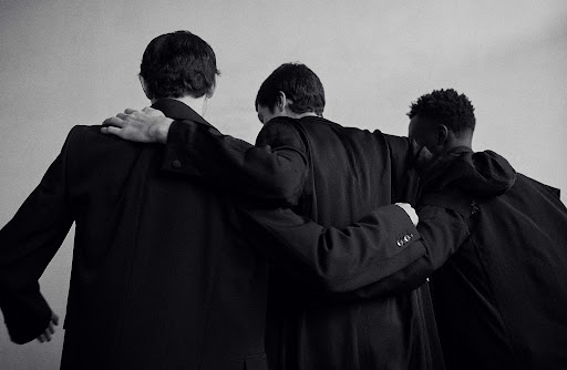 Black and white image of three men in dark jackets or suits standing together with their arms around each other's shoulders.