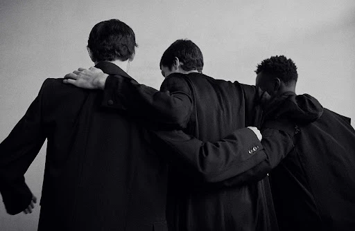Black and white image of three men in dark jackets or suits standing together with their arms around each other's shoulders.