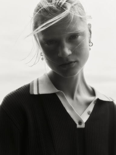 Monochrome portrait of a young woman with blonde hair blowing across her face, wearing a dark polo shirt with a light collar.