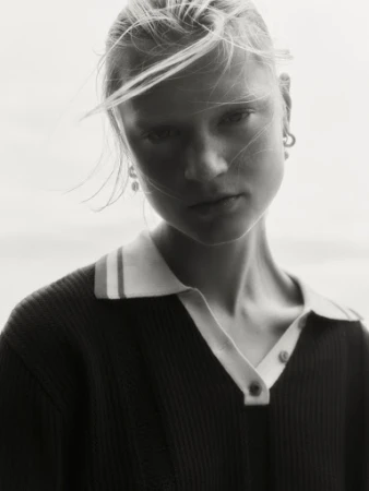 Monochrome portrait of a young woman with blonde hair blowing across her face, wearing a dark polo shirt with a light collar.