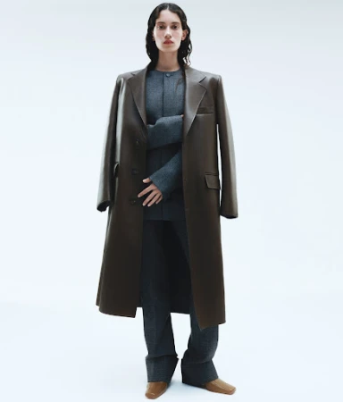 Model wearing a dark leather trench coat and grey suit with styled hair against a plain white background