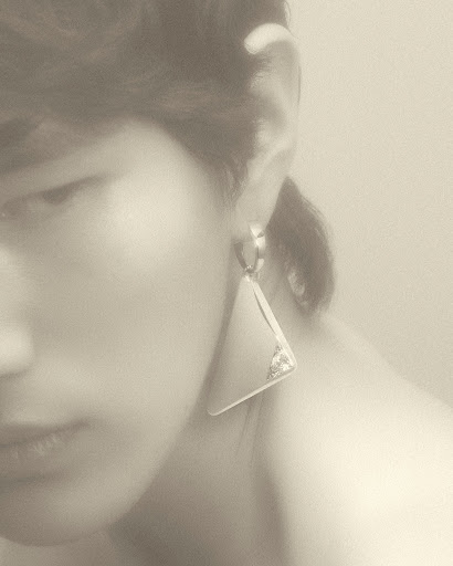 Close up of a woman with fair skin wearing a silver, triangular statement earring. The background is blurred and a neutral tone.