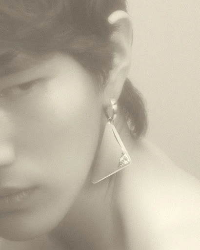 Close up of a woman with fair skin wearing a silver, triangular statement earring. The background is blurred and a neutral tone.