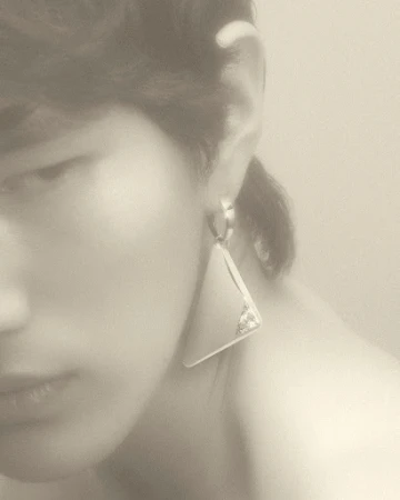 Close up of a woman with fair skin wearing a silver, triangular statement earring. The background is blurred and a neutral tone.