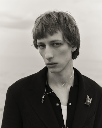 Monochrome portrait of a slender young adult male with a pensive expression, wearing a black jacket, jewelry, against a neutral background.