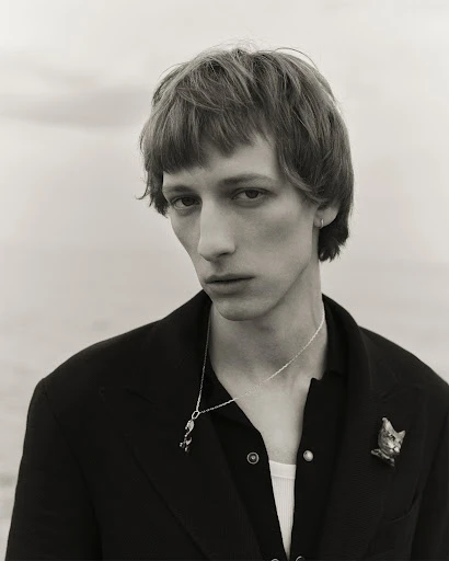 Monochrome portrait of a slender young adult male with a pensive expression, wearing a black jacket, jewelry, against a neutral background.