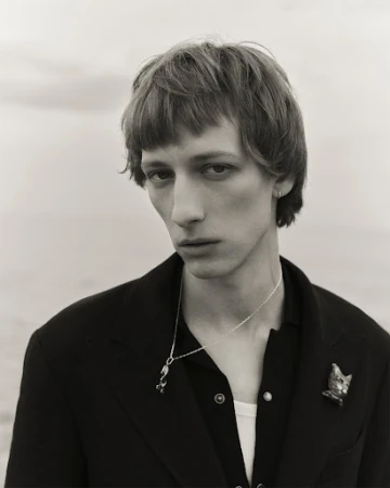 Monochrome portrait of a slender young adult male with a pensive expression, wearing a black jacket, jewelry, against a neutral background.