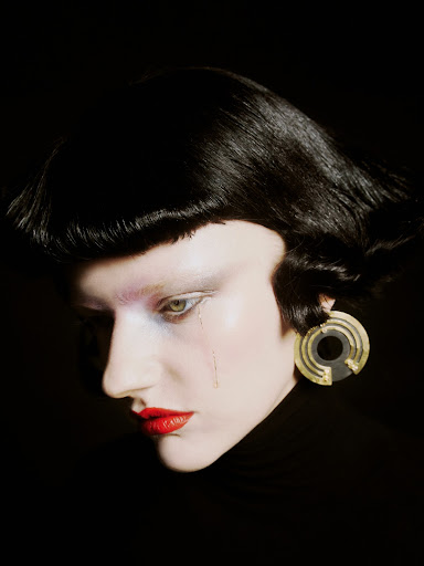 Close-up profile of a model with short black hair, red lipstick, light skin, and a geometric earring against a dark background.
