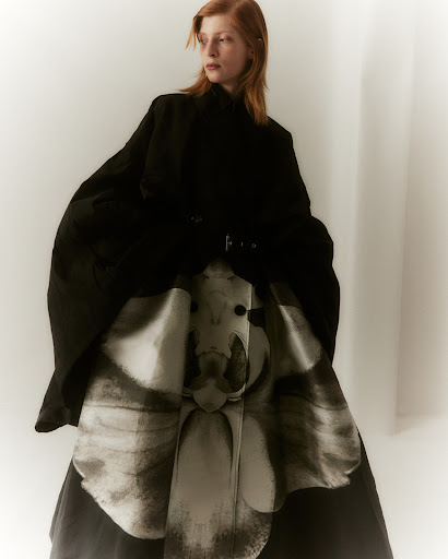 A red-haired fashion model poses in an indoor studio wearing a black jacket and a high-waisted patterned skirt. The shot is a medium studio shot.
