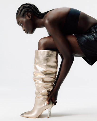 A woman with dark skin and cornrows is wearing a black top, leather skirt, and a gold knee-high boot in a fashion shot.