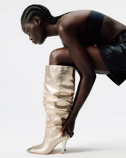 A woman with dark skin and cornrows is wearing a black top, leather skirt, and a gold knee-high boot in a fashion shot.