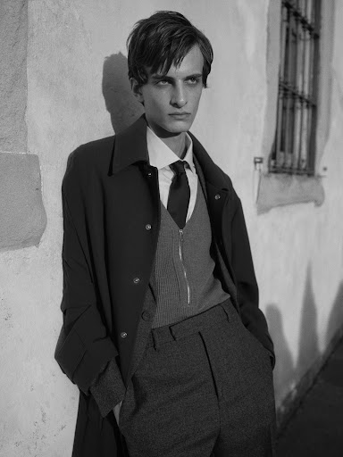 Monochrome fashion portrait of a slender adult male in a coat, sweater vest, and trousers, leaning against a textured wall with a window.