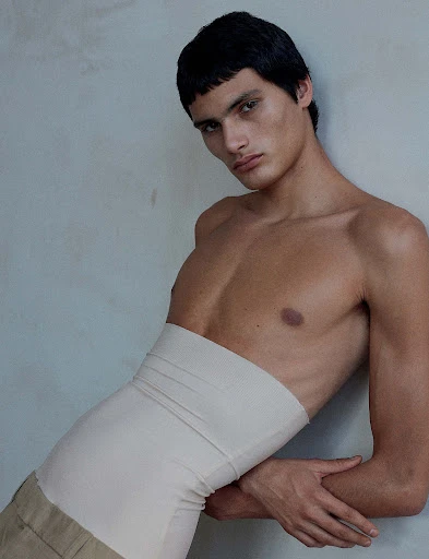 A single, topless male model with dark hair poses indoors against a neutral wall, wearing low beige pants and a waist-high beige elastic bandage.