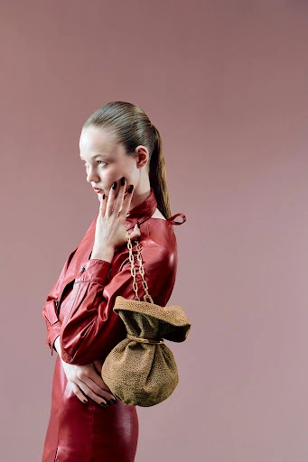A woman in a red dress poses in a photo studio with a tan, drawstring bag. The image has a light pink background.