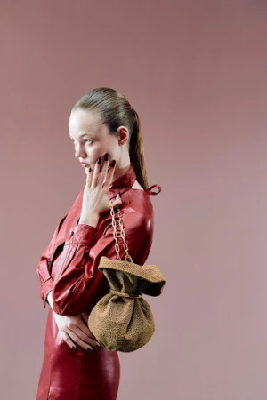 A woman in a red dress poses in a photo studio with a tan, drawstring bag. The image has a light pink background.
