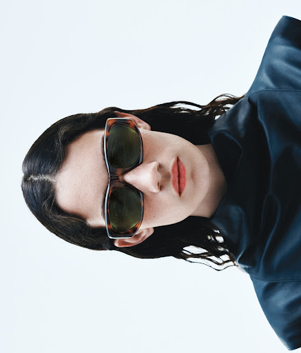 A woman poses in a studio wearing sunglasses and a dark blue leather jacket, set against a plain white backdrop. Fashion shot.
