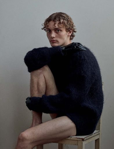 Male model poses on a stool with curly hair, wearing a dark fuzzy sweater and black gloves in a simple, indoor studio setting.