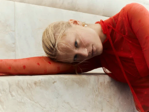 Woman in a red sheer top rests on marble steps, blonde hair partially obscures her face in a tilted close-up, soft natural lighting.