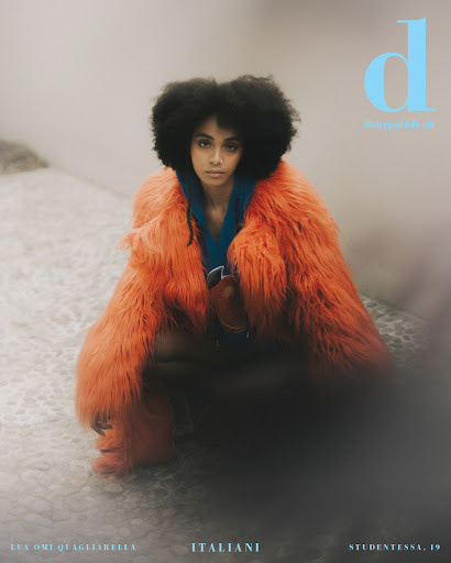 Magazine cover featuring a young woman with afro hair, wearing a furry orange coat, squatting on a patterned surface, contrasted by grey tones.