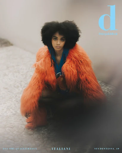 Magazine cover featuring a young woman with afro hair, wearing a furry orange coat, squatting on a patterned surface, contrasted by grey tones.