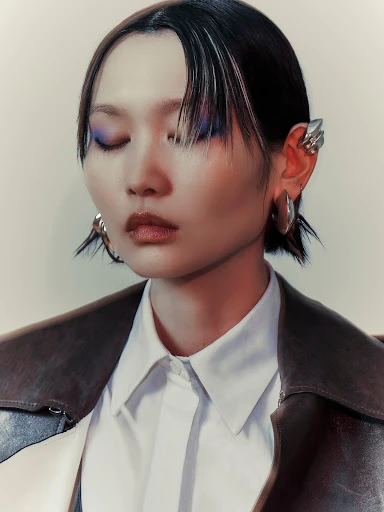 Close-up studio portrait of an Asian woman with closed eyes, wearing artistic silver ear cuffs and a white collared shirt under a jacket.