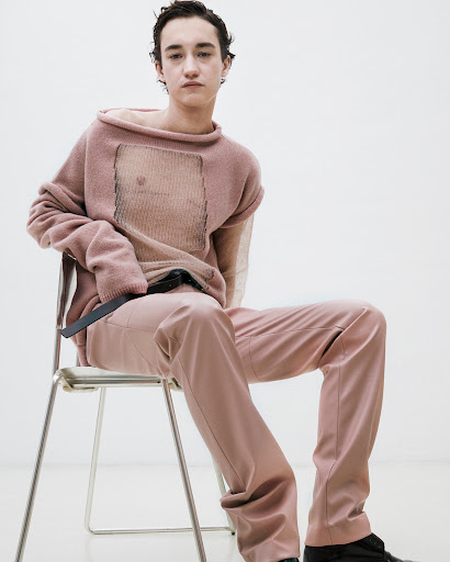 A young man seated on a metal chair wearing a loose dusty pink knit sweater and matching pink trousers against a plain white wall