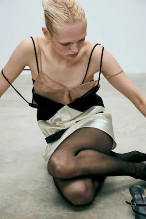 Model seated on the floor wearing a layered black and cream dress with sheer pink straps and black tights