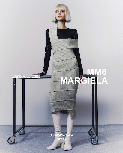 Fashion-forward model poses in a stylish grey knit dress with white boots, standing beside a black table in a stark studio setting.