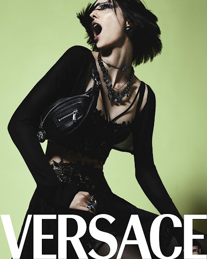 A woman with dark hair poses in a black outfit and jewelry in a studio with a lime green background. The Versace logo is visible.
